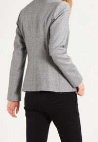 Gray fitted blazer with a subtle houndstooth pattern, structured shoulders, and a single vent at the back, paired with black trousers.