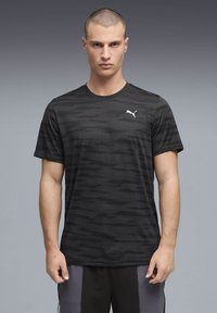 Άνδρας με κοντά μαλλιά, φοράει μαύρο μπλουζάκι Puma με σχέδιο και μαύρο-γκρι σορτς, στέκεται μπροστά σε γκρι διαβαθμισμένο φόντο.