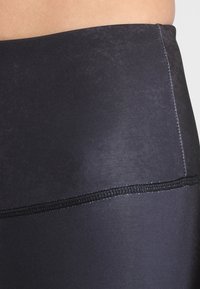 Czarne legginsy o gładkiej, matowej teksturze, z szerokim pasem i kontrastowym przeszyciem wzdłuż szwów.