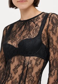 Čierny čipkovaný body s intricnými kvetinovými vzormi, priesvitnou látkou a štruktúrovaným čiernym podprsenkovým košíčkom viditeľným pod ním.
