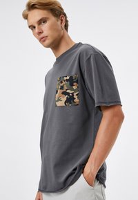 Camiseta de algodón gris con un bolsillo en el pecho que presenta un patrón de camuflaje. Manga corta, corte holgado y detalles de borde sin rematar en el dobladillo.
