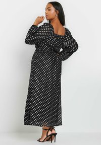 Mustapilkkuinen maksi mekko, jossa on läpinäkyvät pitkät hihat, vyötäröltä kiristetty muotoilu ja neliönmallinen kaula-aukko, yhdistettynä mustiin korkokenkiin.