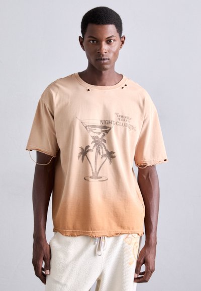 Man in een vervaagde perzikkleurige T-shirt met een palmboom- en cocktailglasafbeelding en witte gestructureerde broek, staand tegen een effen lichte muur.