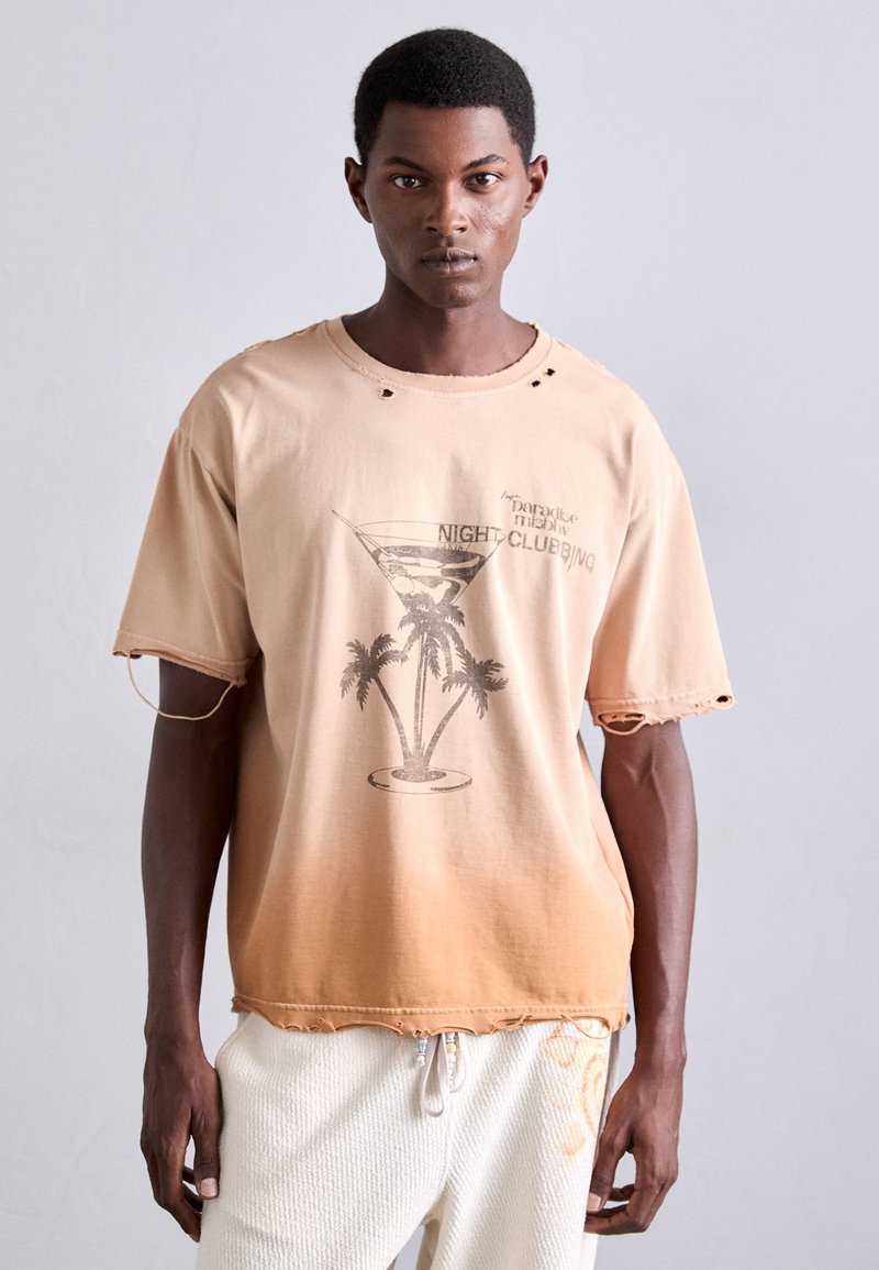 Mand iført en falmet ferskenfarvet t-shirt med grafik af palmetræ og cocktailglas samt hvide teksturerede bukser, stående op ad en ensfarvet lys væg.