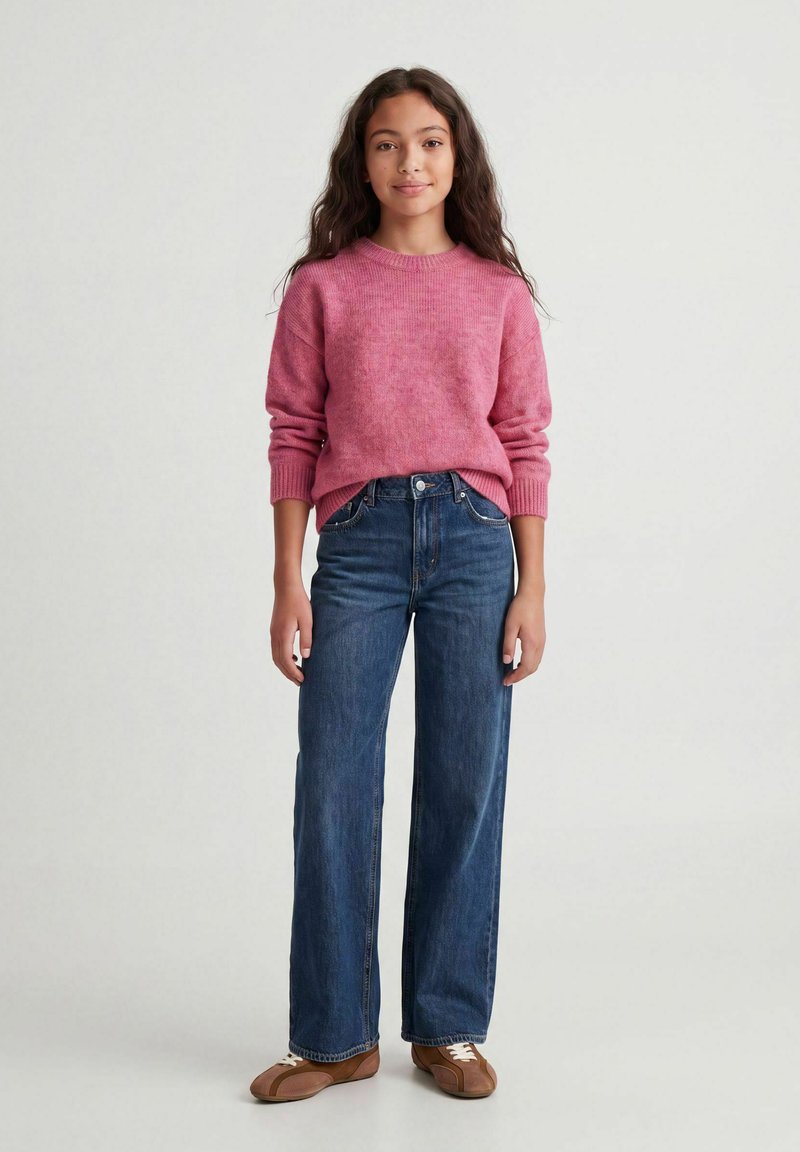 Fille aux longs cheveux ondulés portant un pull rose, un jean bleu à jambes larges et des chaussures marron à lacets, debout devant un fond clair uni.