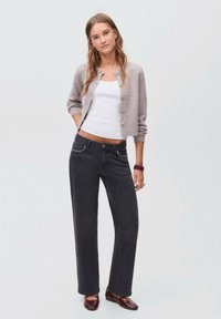 Cardigan grigio chiaro con bottoni, canotta bianca e jeans a gamba larga grigio scuro con dettagli in borchie. Pantofole marroni completano l'outfit.