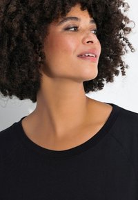 Camiseta negra de cuello redondo con mangas raglán, textura suave y detalles de costura mínimos. El modelo tiene el cabello rizado y maquillaje neutro.