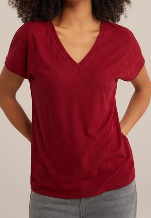 Chemise rouge à manches courtes avec col en V et motif texturé en forme de feuille. Tissu doux, coupe décontractée, et détail d'ourlet arrondi. Associée à un jean gris.