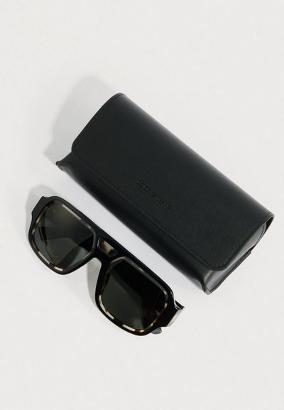 Μαύρα γυαλιά ηλίου με σχέδιο χελώνας και σκοτεινούς φακούς δίπλα σε μαύρη ορθογώνια δερμάτινη θήκη Dolce & Gabbana, πάνω σε λευκή επιφάνεια.