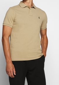 Beige polo skjorta tillverkad av mjuk bomull med klassisk krage, korta ärmar och en liten grön logotyp på vänster bröst.