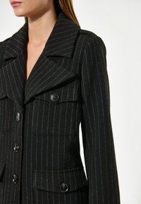 Črno-zlata blazer iz volne s črtami, z velikimi reverji, dvojno zapiranje z gumbi in dvema sprednjima žepoma. Teksture barve.