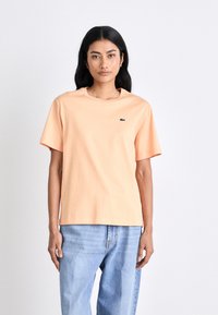 Pfirsichfarbenes T-Shirt mit kurzen Ärmeln aus Baumwolle, Rundhalsausschnitt und grünen Logodetails, kombiniert mit hellblauen Jeans. Schlichte, lockere Passform.