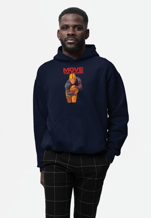 Mann trägt einen marineblauen Hoodie mit einer Vintage-Fußballspielergrafik und dem roten Text „MOVE THE CHAINS“, kombiniert mit schwarz karierten Hosen.