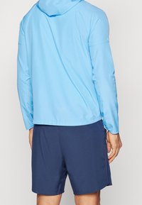 Veste légère à capuche bleu clair à manches longues, associée à un short bleu marine foncé ; présente une texture lisse et un design décontracté.