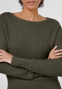 Pull en laine vert olive texturé avec un col bateau, présentant un motif ondulé, des poignets côtelés et une coupe ajustée.