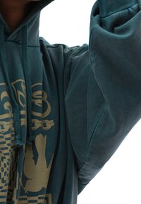 Hoodie teal realizzato in tessuto morbido, con un design grafico dorato che presenta motivi di catene e un uccello. Lacci con frange nel cappuccio.