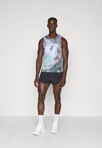Ärmelloses Sportoberteil für Herren im mehrfarbigen Muster mit abstraktem Design; kombiniert mit schwarzen Shorts und weißen Turnschuhen. Glatte Stofftextur.