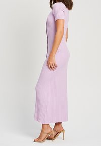 Femme portant une robe longue en maille côtelée, à manches courtes, de couleur violet clair, avec des sandales à talons hauts beige, debout de profil sur un fond uni.