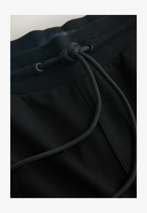 Mustad pükstega joggerid, millel on nööriga vöökohad ja tekstuuriga kangas, rõhutades sujuvaid jooni ja minimalistlikku disaini.