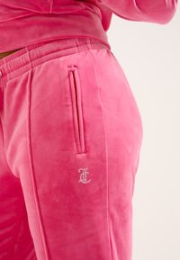 Roza žametne trenirka hlače s stranskim žepom na zadrgo in okrašenim logotipom iz kristalov. Mehka tekstura, prilegajoča zasnova in elastičen pas.
