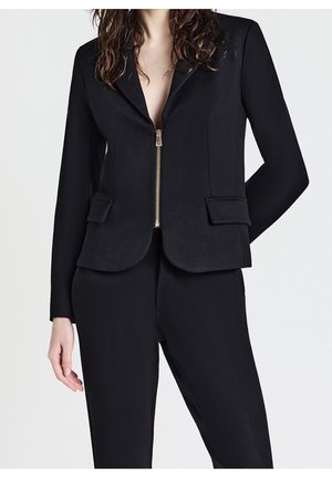 Kvinde iført en tætsiddende sort blazer med lynlås foran og klaplommer, kombineret med matchende sorte bukser mod en ensfarvet baggrund.