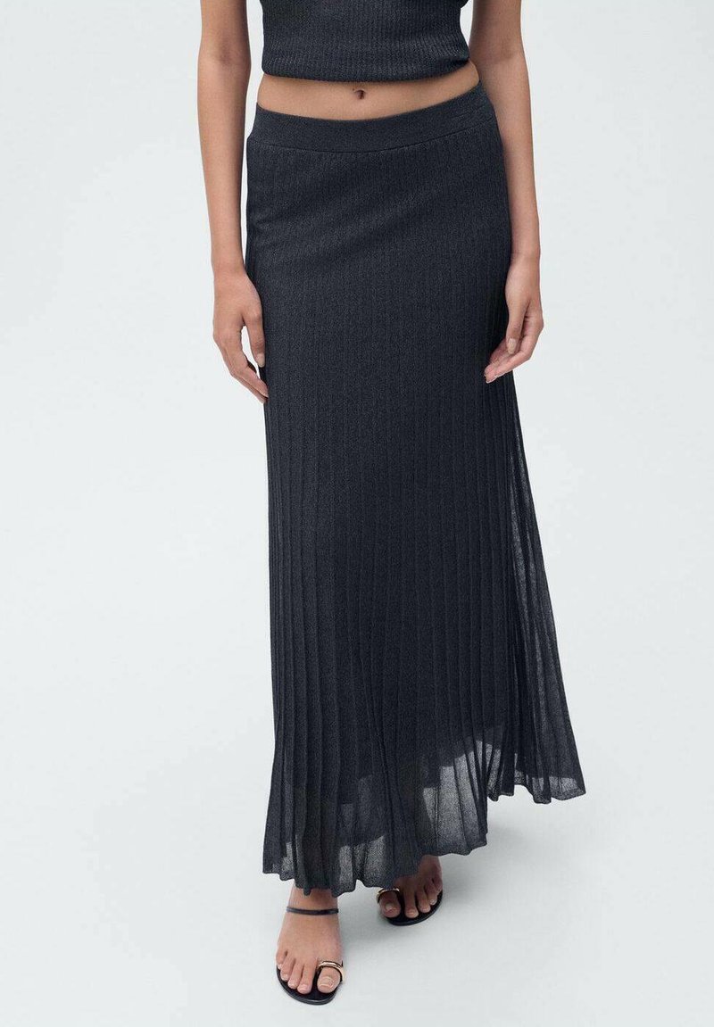 Jupe plissée longue et gris foncé en tissu léger. Texture lisse avec une taille large, associée à des sandales noires laissant voir les orteils.