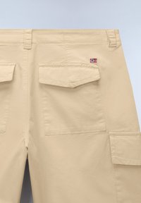 Pantalones cargo beige con bolsillos con solapa, trabillas para cinturón y un pequeño parche con la bandera noruega encima del bolsillo trasero derecho.