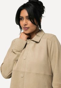 Beige Wildlederjacke mit Kragen, Knopfverschluss und glatter Textur. Besitzt sichtbare Nähte und ein entspanntes Design.