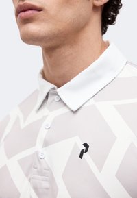 Κοντινό πλάνο ενός ατόμου που φοράει λευκό πόλο μπλουζάκι με γεωμετρικό σχέδιο και μικρό μαύρο λογότυπο στο στήθος.