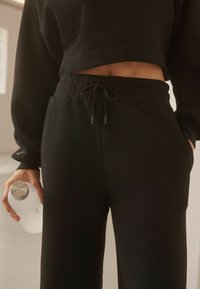Sort cropped sweatshirt med elastisk talje og joggers med justerbar snøre og sidelommer. Modellen holder en hvid vandflaske.