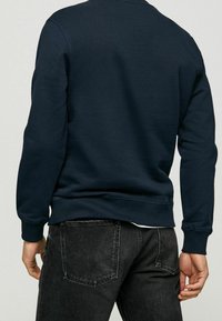 Mörkblå tjocktröja i bomullsblandning med strukturerad yta. Har ribbade muddar och nederkant, bärs med svarta denimjeans.