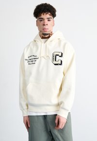 Sudadera de color crema con un bolsillo frontal tipo pouch, capucha con cordón y detalles gráficos en negro que incluyen texto y una letra "C" en el pecho.