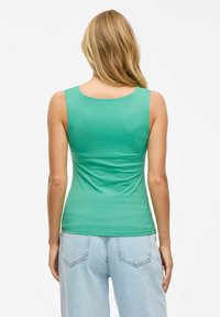 Zelena brezrokavna majica iz gladkega, elastičnega materiala s širokim izrezom in nabrano obdelavo na hrbtni strani, v kombinaciji z svetlo modrimi kavbojkami.