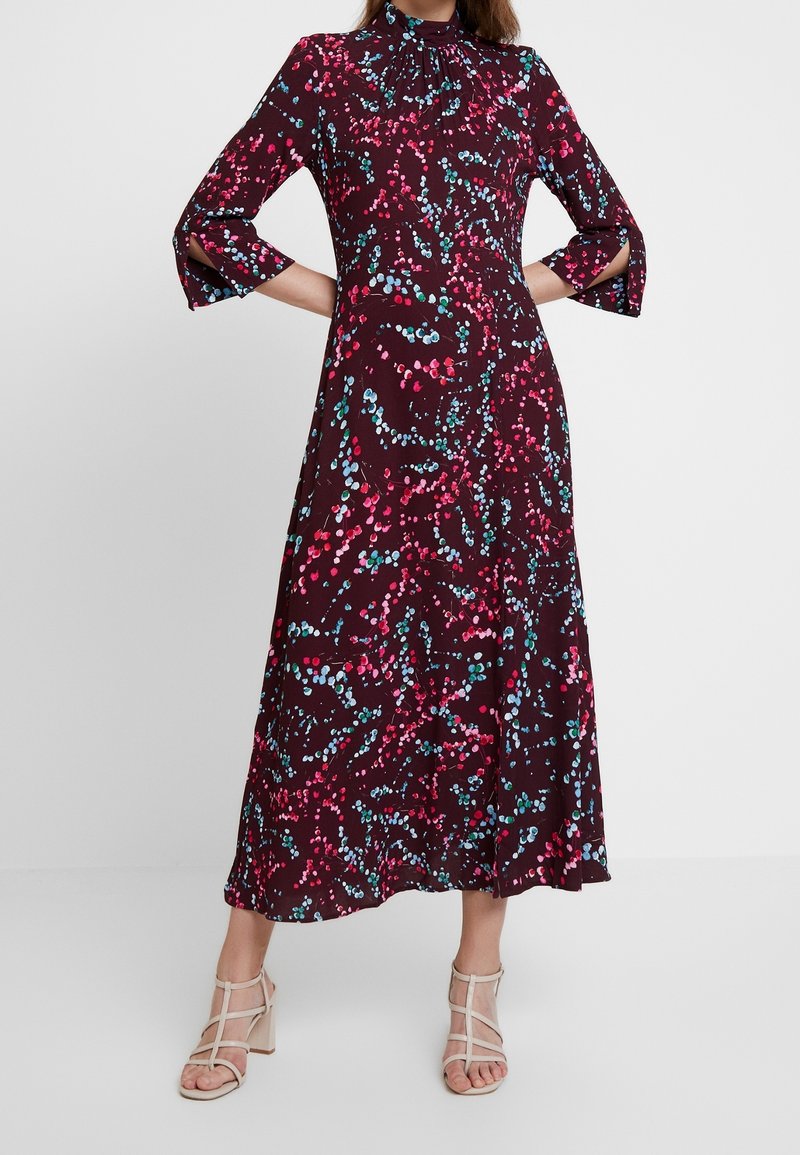 Femme portant une longue robe florale sombre avec des motifs à pois roses et bleus, des manches fendues aux trois-quarts, et des talons ouverts beige.