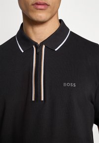 Polo noir avec un col zippé orné de rayures contrastantes blanches et beiges. Le nom de la marque "BOSS" est discrètement affiché sur la poitrine.