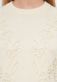 Pull en maille couleur crème avec des motifs floraux en relief brodés sur le devant et un col rond.