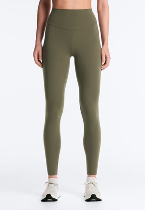 Vue de face d'une personne portant des leggings taille haute vert olive et des chaussures de sport blanches, debout avec les bras détendus le long du corps.