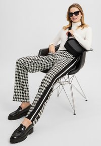 Woman wearing white turtleneck and black-and-white houndstooth pants with side stripes, sitting on black chair, holding black crossbody bag.