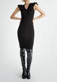 Robe noire ajustée avec manches à volants, décolleté en V, coutures structurées. Associée à des bottes noires vernies montantes jusqu'aux cuisses, texture lisse et brillante.