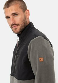 Fleecejacke mit schwarzem Oberstoff, grauem Fleece-Unterteil, hohem Kragen, Reißverschluss-Taschen auf der Vorderseite und einem Leder-Logo-Patch am Ärmel.