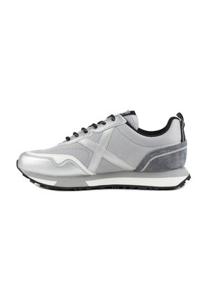 Zapatilla deportiva gris y plateada con cordones negros, suela blanca y banda de rodadura negra vista de perfil lateral sobre fondo blanco.