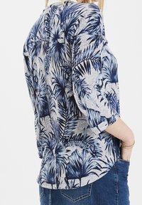 Grijze top met een blauw palmbladpatroon, driedelige mouwen en een ronde zoom; gemaakt van zachte stof met een ontspannen pasvorm.