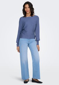 Pull côtelé bleu avec un large col, associé à un jean taille haute bleu clair et des ballerines noires, sur un fond blanc uni.