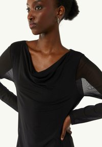 Top noir drapé avec des manches longues transparentes et un col en cowl. Tissu doux avec une texture subtile, design ajusté et surface lisse.