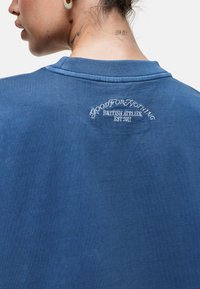 Blauwe sweater van zacht materiaal. Heeft een geribbelde kraag en geborduurde tekst op de achterkant: "GOOD FOR NOTHING BRITISH ATELIER EST. 2011."