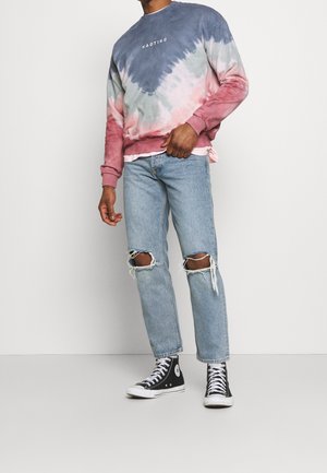 Sudadera de tie-dye en tonos azules, rosa y blanco con puños acanalados; combinada con vaqueros claros rotos y zapatillas altas negras.