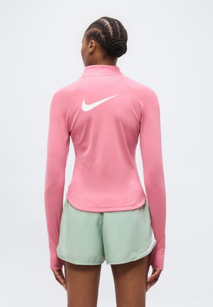 Ženska s spletenimi lasmi, ki nosi oprijet svetlo roza dolg rokav s belim logotipom Nike in svetlo zelenimi športnimi kratkimi hlačami, stoji obrnjena stran.