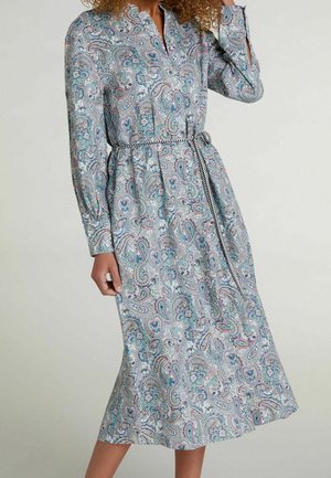 Femme portant une robe à manches longues ceinturée avec un motif paisley dans des tons de bleu, vert et violet, debout devant un fond uni.