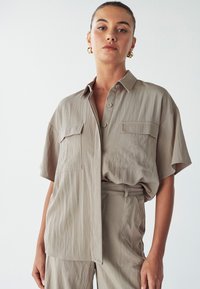 Camicia a maniche corte color beige chiaro con vestibilità rilassata, due tasche sul petto e chiusura con bottoni. Il tessuto ha un aspetto leggermente testurizzato.