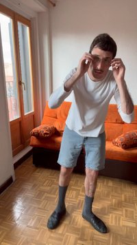 Mann i hvit langermet skjorte og denimshorts justerer brillene sine, stående på parkettgulv nær oransje sofa og vindu.